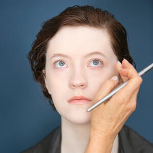 Ky Mullen, a white androgynous person with auburn hair looks up past the camera. There's a hand with a makeup brush resting on their cheek.